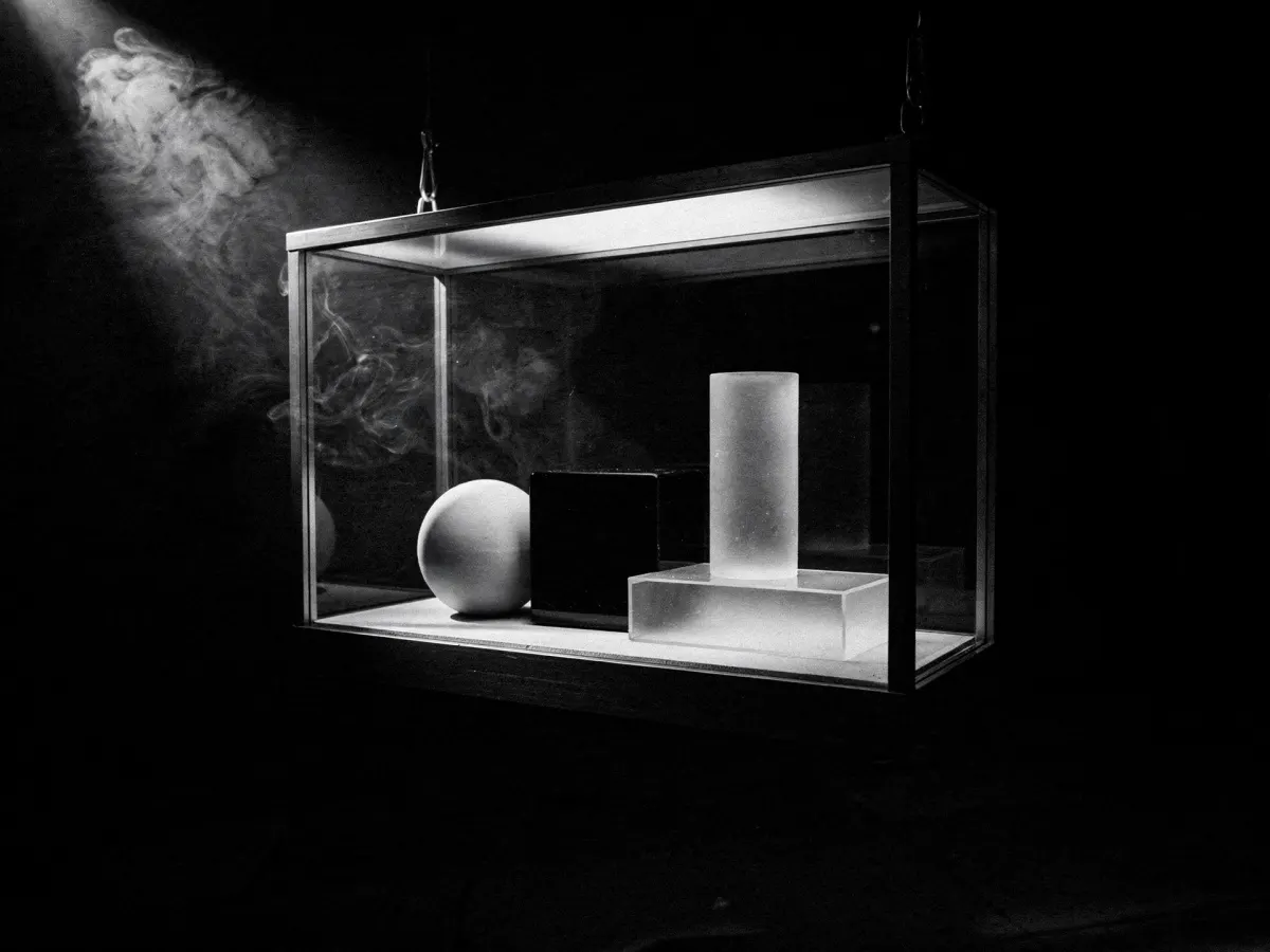 A lit glass display case holding simple geometric shapes, floating in a black void