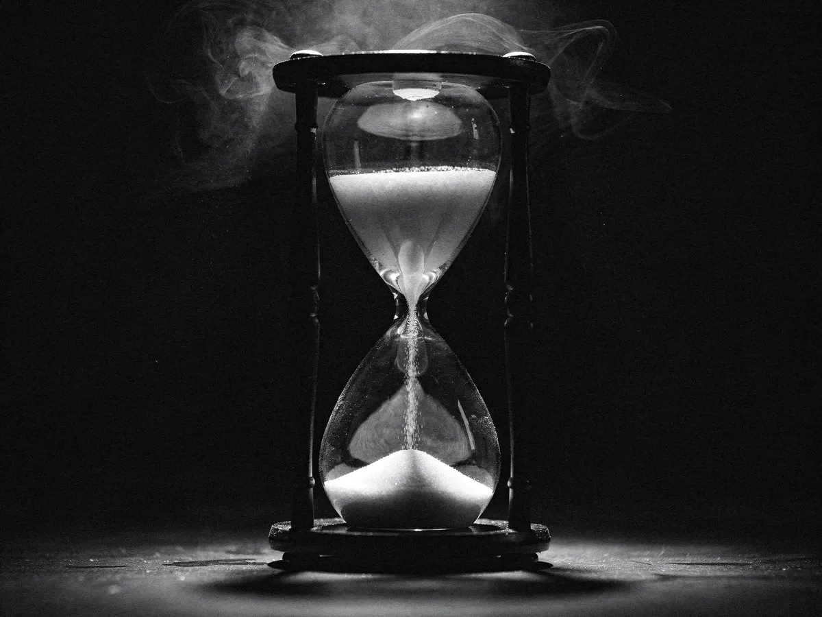 A single hourglass in a dark wooden frame, centred under a hard light, the sand mid-fall
